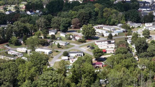 Rows of single wide manufactured homes sit along curving drives in a wooded mobile home park at Lynchburg, Virginia, late summer canopy closing in around the lots. Aerial drone wide shot