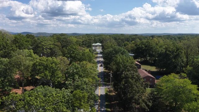 A two lane residential road runs between mature oaks toward a white water tower at Lynchburg, Virginia, brick homes and apartment blocks set back among the trees. Aerial forward wide shot