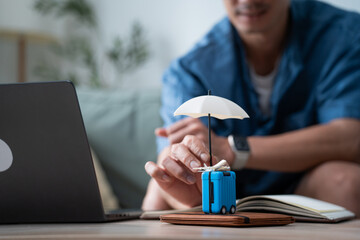 A man plans a vacation by holding a small luggage model protected by a tiny umbrella next to a laptop at home, conveying themes of travel insurance, financial planning, and preparation. © winnievinzence