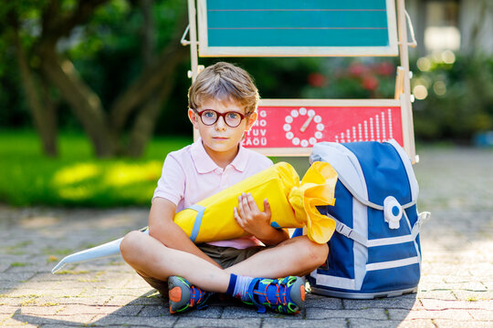 Happy little kid boy with glasses sitting by desk and backpack or satchel. Schoolkid with traditional German school bag cone called Schultuete on his first day to school