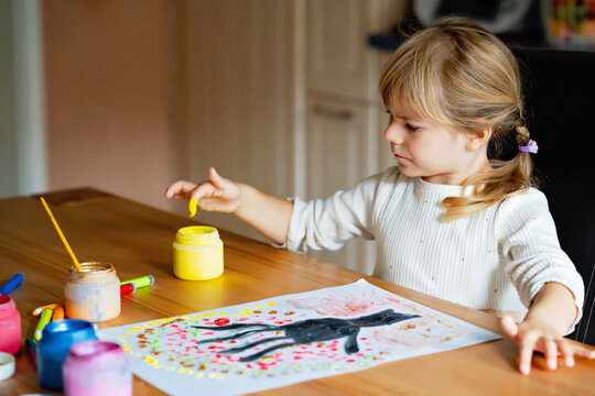 Little preschool girl painting with fingers tree on paper at home or at kindergarden daycare. Child doing creative art activity indoors. Real life, real people.