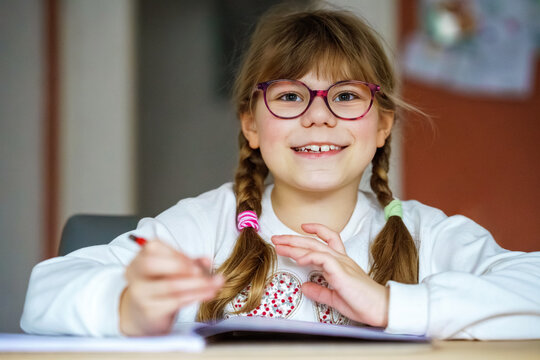 Smiling school girl with glasses writing in notebook at desk, happy child studying at home, education and learning concept. Elementary school child doing homework.