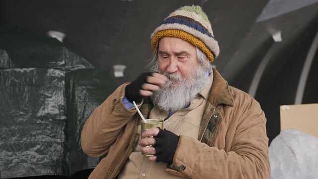 Senior Caucasian homeless man eating canned food with plastic spoon outdoors, wiping mouth. Ideal for social welfare campaigns, charity food drive commercials, poverty awareness videos