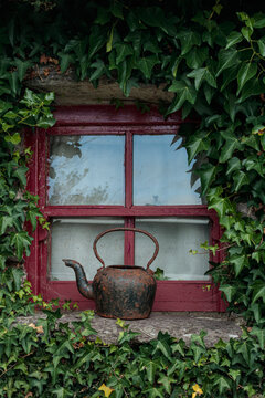 Old rusty iron kettle sitting on a windowsill framed by red wooden window frame and lush green ivy leaves