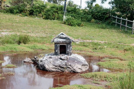 Small miniature stone house model standing on a large rock in the middle of a shallow pond surrounded by green grass