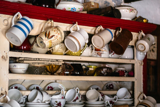 Collection of retro tea cups, saucers, and ceramic mugs hanging on hooks in traditional kitchen dresser