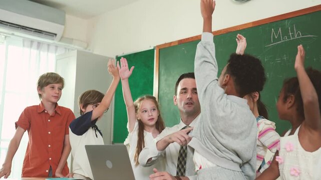 Elementary students raise hands to answer teacher during technology lesson with laptop at front of classroom, showing digital learning, participation, computer education.