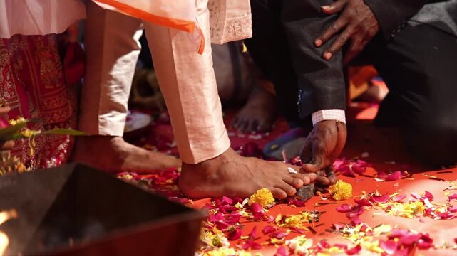 A close-up, low-angle shot of bare feet, likely belonging to a groom in traditional Indian attire, stepping onto a vibrant red floor adorned with scattered red and yellow flower petals, indian wedding