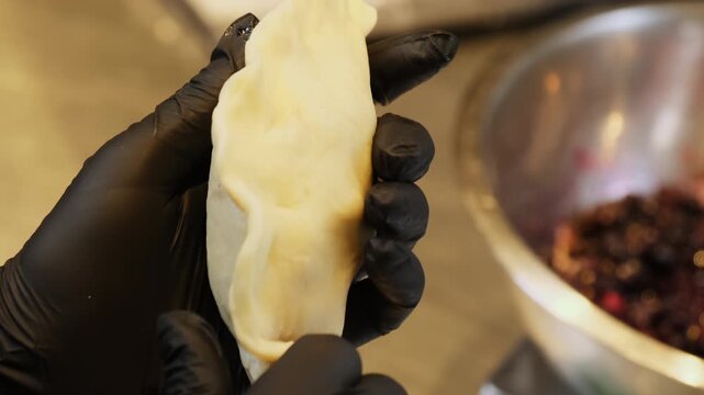 Gloved baker seals filled dumpling edges by hand near bowl of berry filling. Fingers press soft dough together closing pastry securely