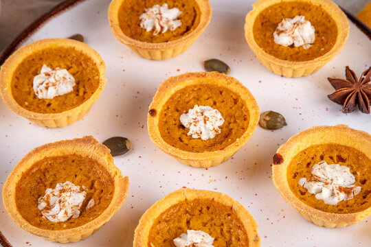 Sweet autumn baking, Mini pumpkin Tarts. Tiny pumpkin spice pies, with whipped cream topping and cinnamon powder, on beige concrete background