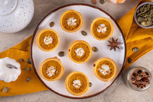 Sweet autumn baking, Mini pumpkin Tarts. Tiny pumpkin spice pies, with whipped cream topping and cinnamon powder, on beige concrete background