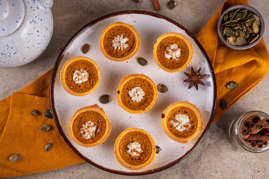 Sweet autumn baking, Mini pumpkin Tarts. Tiny pumpkin spice pies, with whipped cream topping and cinnamon powder, on beige concrete background