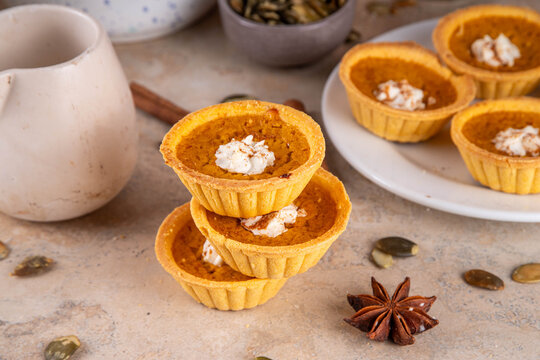 Sweet autumn baking, Mini pumpkin Tarts. Tiny pumpkin spice pies, with whipped cream topping and cinnamon powder, on beige concrete background