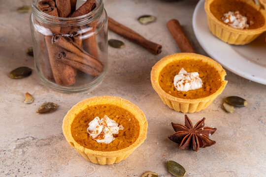 Sweet autumn baking, Mini pumpkin Tarts. Tiny pumpkin spice pies, with whipped cream topping and cinnamon powder, on beige concrete background