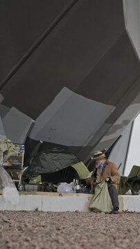 Vertical shot of senior Caucasian homeless man sitting near makeshift shelter sorting belongings. Clip suitable for poverty awareness campaigns, charity documentaries and social aid videos