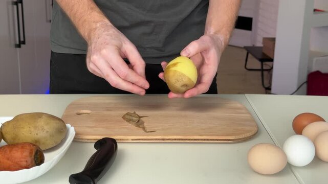 Hands peeling yellow potato with peeler over wooden cutting board in bright modern kitchen, close-up fingers shaping potato skin, food preparation scene, countertop work surface view