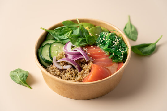 Healthy protein bowl with quinoa, smoked salmon, spinach, cucumber, avocado and chuka seaweed salad in a cardboard container on a light beige background