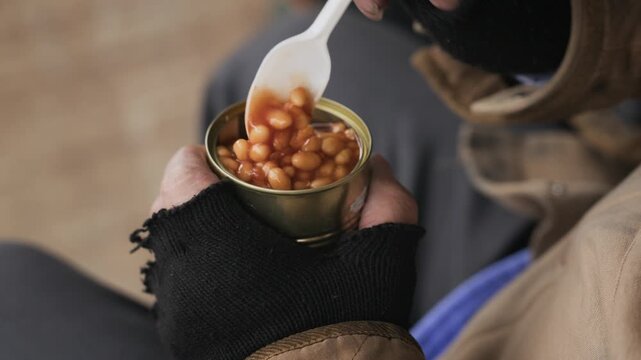 Homeless man eating canned beans from tin with plastic spoon. Ideal for social welfare campaigns, poverty awareness videos, and hunger relief charity projects