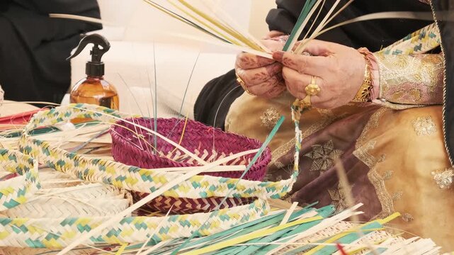 Skilled hands weave palm frond strips into basket form with careful rhythm. Gold jewelry and embroidered dress suggest Emirati heritage, while colored fibers fan outward in traditional Arab craft.