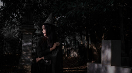 A mysterious witch in a black costume and pointed hat stands in a dark cemetery, surrounded by tombstones and deep shadows, creating an eerie Halloween atmosphere. © Kieng