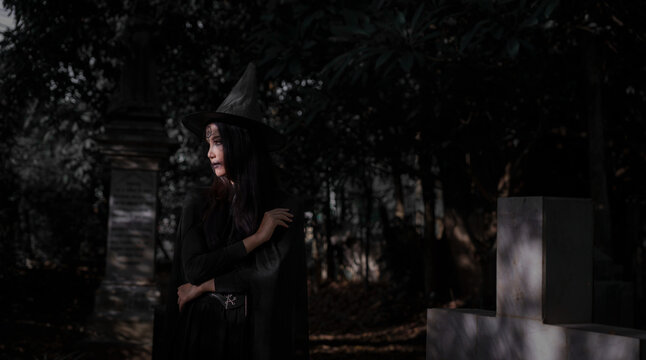 A mysterious witch in a black costume and pointed hat stands in a dark cemetery, surrounded by tombstones and deep shadows, creating an eerie Halloween atmosphere.