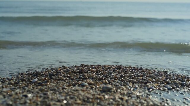 Pebbles beach sand sea waves, handheld static shot with a close-up focus on a small pile of wet pebbles and seashells on the shore with blurred ocean waves breaking in the background during bright sum