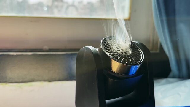 Close-up of a black electronic aroma diffuser emitting white vapor indoors. The portable device sits next to a bright window with a blurred city view during daylight, near a blue curtain.
