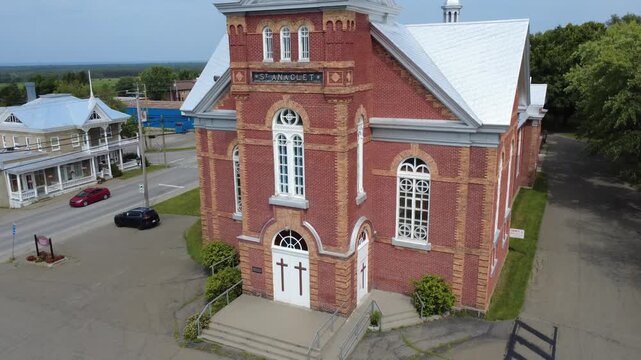 Drone footage approaching the red‑brick Catholic church fa&ccedil;ade in Saint‑Anaclet‑de‑Lessard, showcasing its eclectic architecture and heritage status within Quebec&rsquo;s Bas‑Saint‑Laurent region.