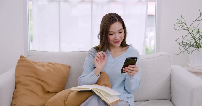 Young Asian woman uses phone while writing daily task note on sofa, then continues planning in notebook, creating calm home productivity moment inside cozy minimal apartment with soft window light