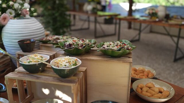Green ceramic bowls of fresh salad and fried snacks on wooden crates at an outdoor catering buffet with flowers in the background.