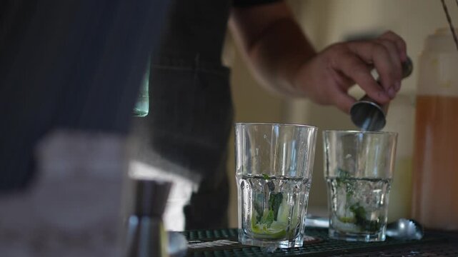 Two tall glasses holding fresh mint and lime wedges stand on a bar counter while a bartender works out of focus behind them.