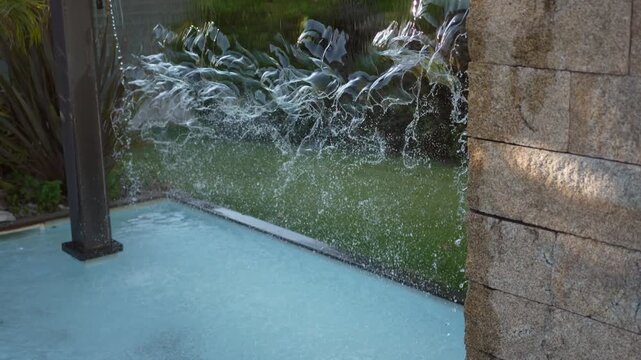 Sheet of water pours from a granite wall and splashes into a turquoise swimming pool at a modern villa on a sunny afternoon.