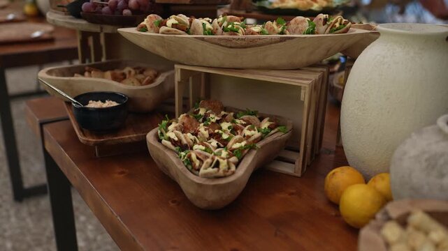 Hand carved wooden bowls filled with salad and fried snacks stand on a dark table beside ceramic pots and lemons at a catering buffet.