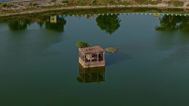 Aerial drone view of a temple teppakulam with a small mandapam standing in the center. Still water reflects the structure and surrounding greenery across the pond.
