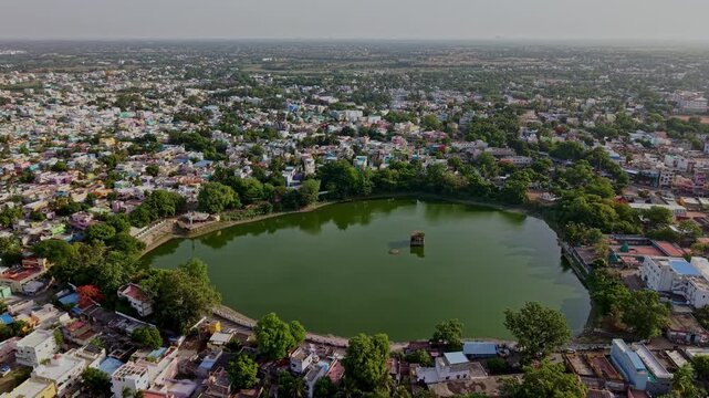 Aerial drone view of a large teppakulam temple tank surrounded by dense urban development. A small mandapam stands at the center of the calm water in Tamil Nadu.