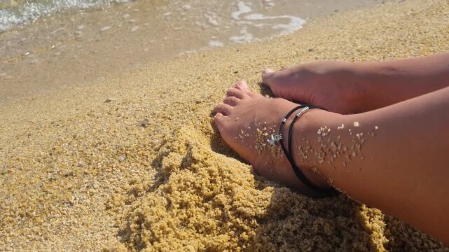 Close-up of a woman&rsquo;s tanned legs and feet with an ankle bracelet resting in soft beach sand, while gentle ocean waves approach her toes, capturing a relaxed summer beach lifestyle