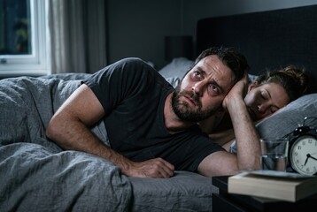 Fototapeta premium A man lies awake in bed with a worried expression. He rests his head on his hand while a woman sleeps beside him. A clock shows the time