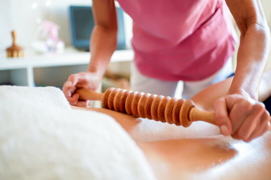 Therapist applying wooden massage roller across bare back of adult client during calm bodywork session in bright treatment room with hands, forearms, table, towels, soft natural light and soothing
