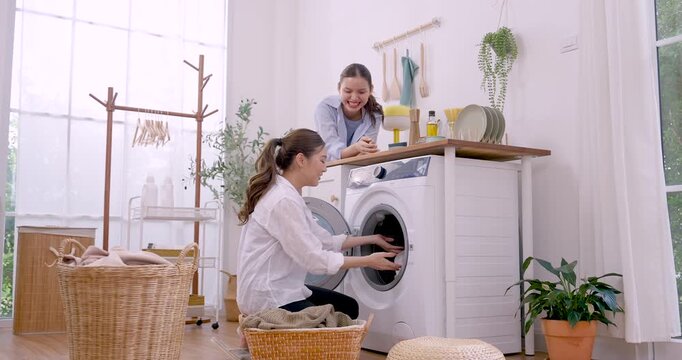 Asian LGBTQ female couple loads clothes into washing machine, passing fabric from basket and reaching into drum near bright window, cozy weekend laundry routine, shared domestic teamwork and support