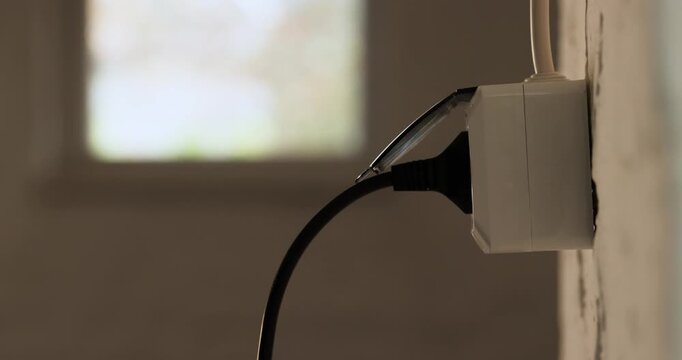 Close-up shot of a male hand inserts a black power cord into a white wall socket indoors with soft natural light.