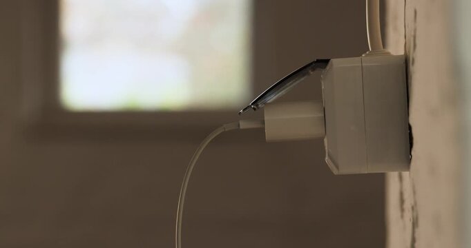 Close-up view of human hand opens a plastic cover and plugs a white modern charger into a wall socket inside a dimly lit garage space.