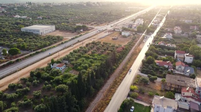 Drone aerial view over a coastal road in Patras, Greece, as the sun sets over the Ionian Sea, revealing warm golden light, urban surroundings and a peaceful Mediterranean setting
