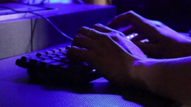 A person types on a computer keyboard at a desk illuminated by blue ambient light to work late at night.