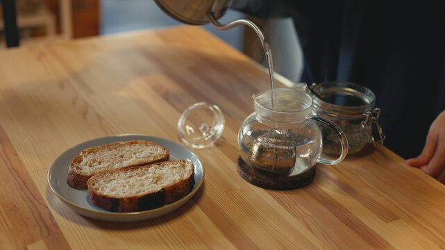 Close-up of person pouring hot water from metal gooseneck kettle into glass teapot with tea strainer. Making tea with sliced bread on wooden table in home kitchen. Morning breakfast routine concept.