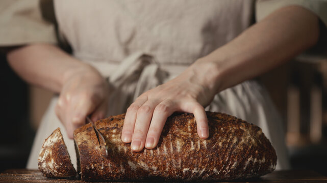 Close-up of baker in rustic linen apron slicing freshly baked artisan sourdough bread with knife on wooden cutting board. Homemade crusty loaf for breakfast, bakery promos, and healthy food concepts.