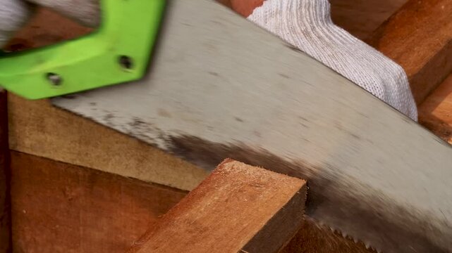 Woodworker saws wooden beam using manual hand saw with green handle in workshop.