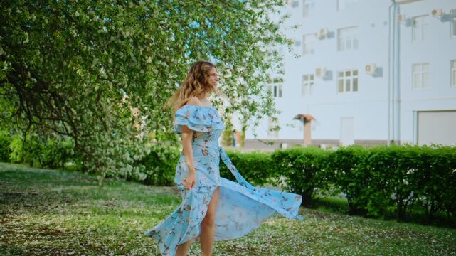 Woman in blue floral dress dances joyfully under blossoming tree. Her arms spread wide, hair flows freely with movement. Expression shows pure happiness and freedom