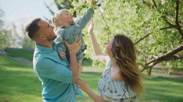 Happy family enjoys a sunny day outdoors. Father holds toddler while mother lifts baby's hand to touch blossoming tree branches. All smile and laugh with joy. Perfect for family, parenting