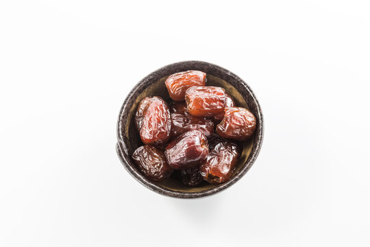 Glossy dried date fruits are arranged in a small dark bowl, isolated on a plain white background. They are a sweet, nutritious dried fruit suitable for snacking or culinary use.