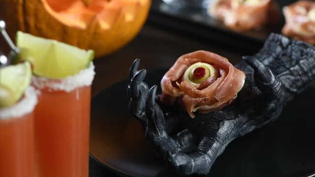 A close-up of a spooky Halloween table setting featuring a bacon and mozzarella eyeball appetizer held by a black skeletal hand on a black plate, amidst drinks and festive decor. Horizontal video, rig